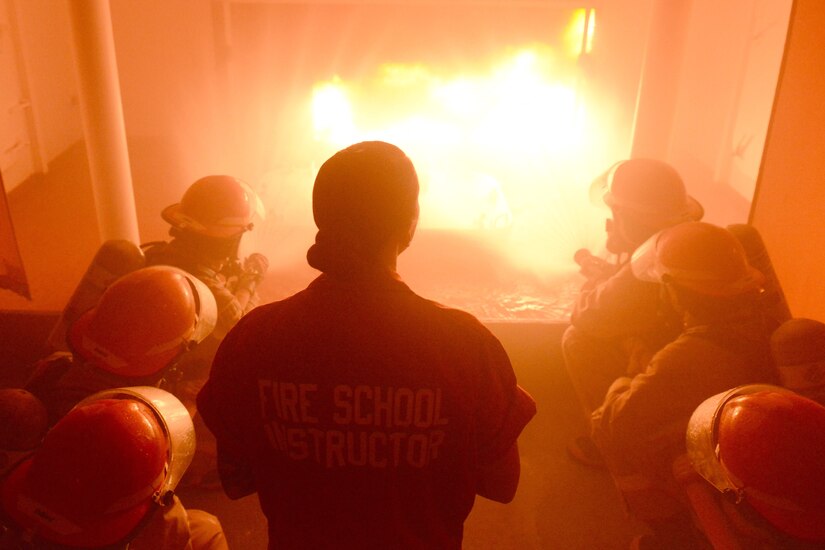 A woman wearing a jumpsuit marked "Fire School Instructor" stands over Coast Guardsmen in fire gear as they aim hoses at a fire.