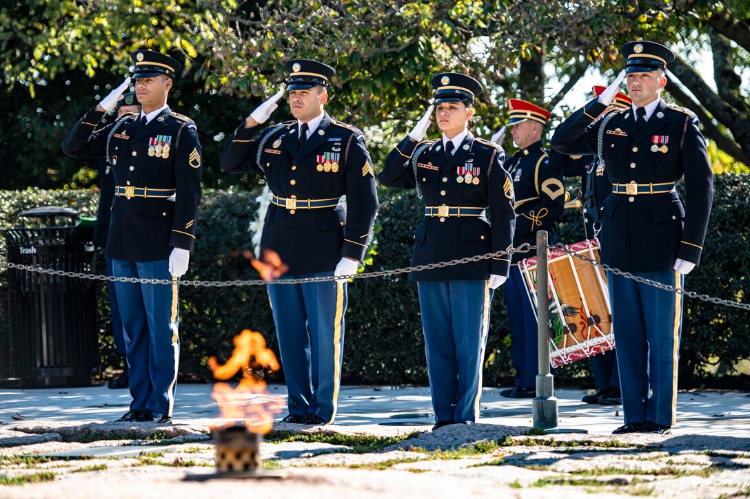 Four soldiers salute in front of a flame at a cemetery.