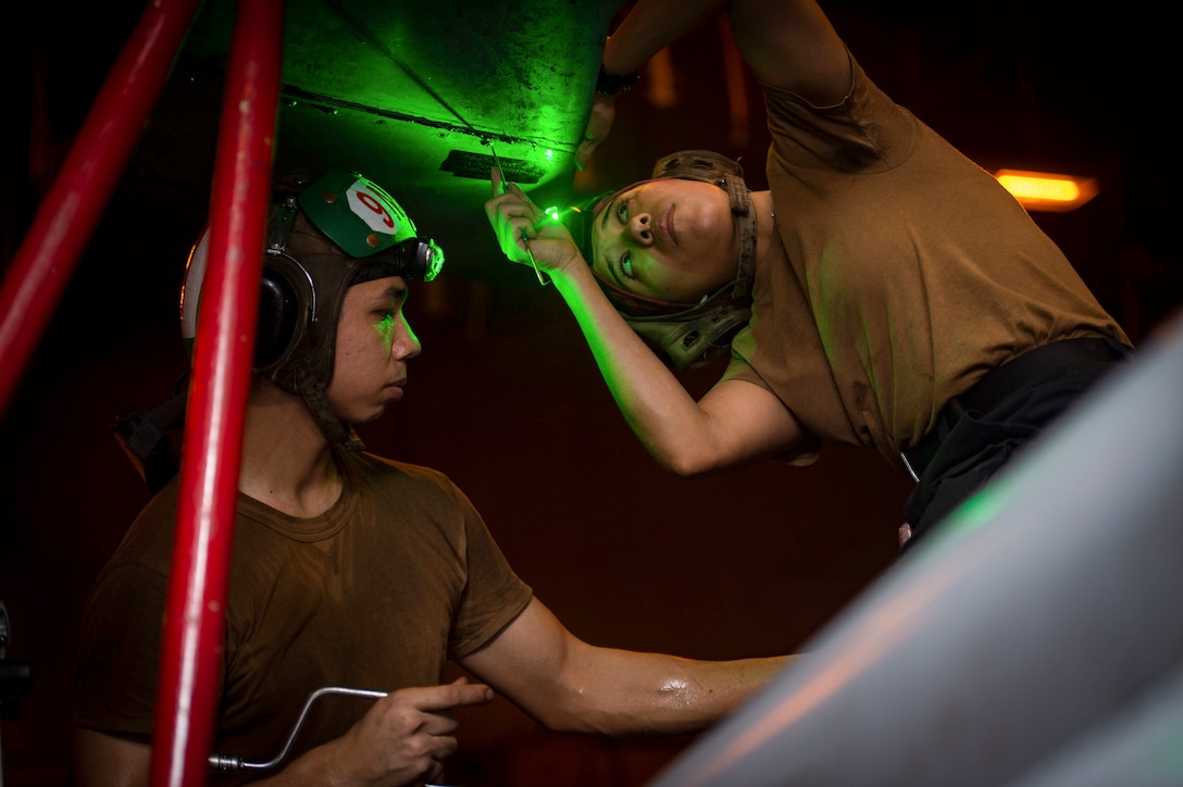 A sailor uses a green light to look up at an aircraft section as another assists.