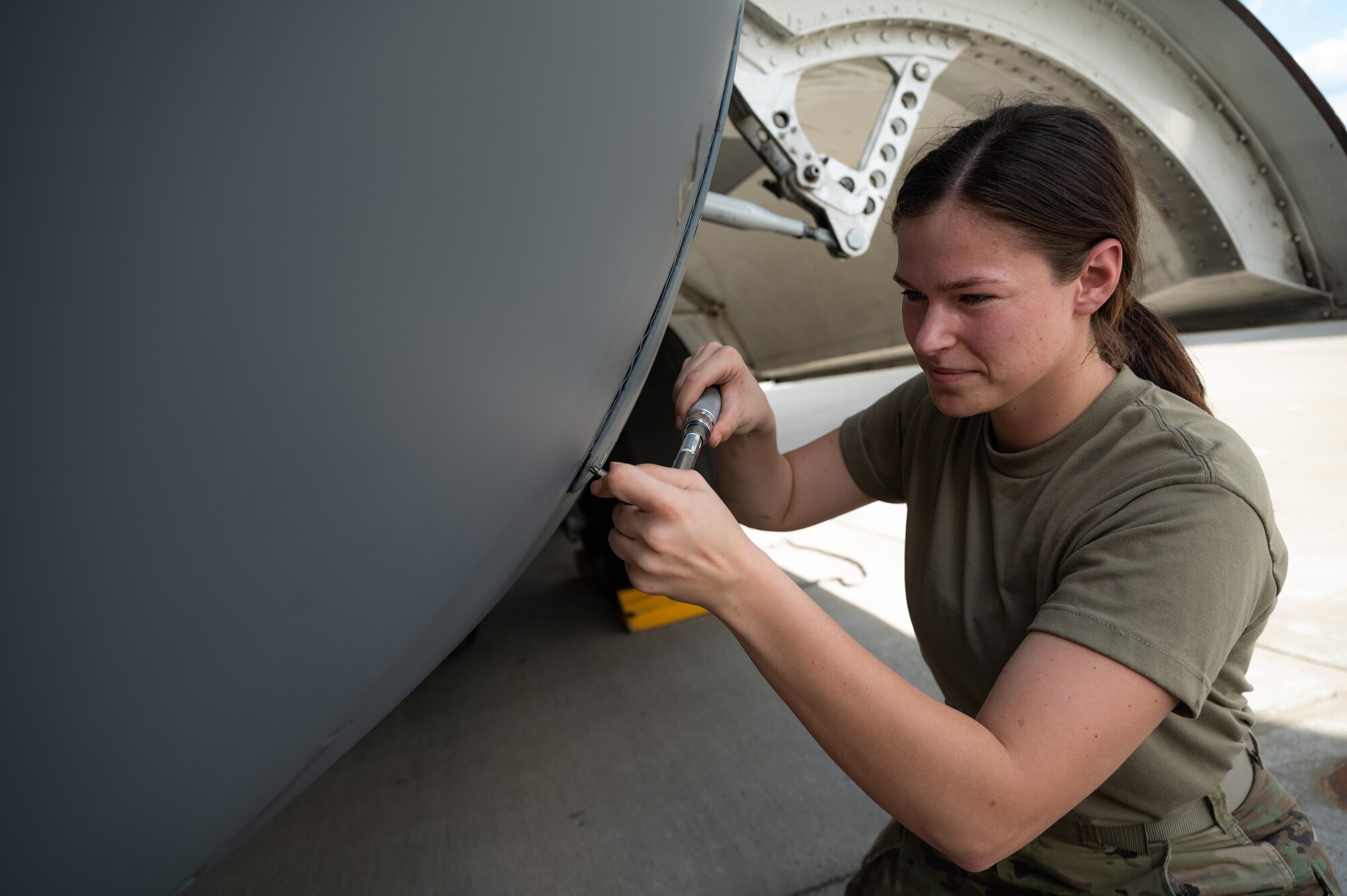 A female Airmen holds a wrench while working on a panel on the side of a large aircraft