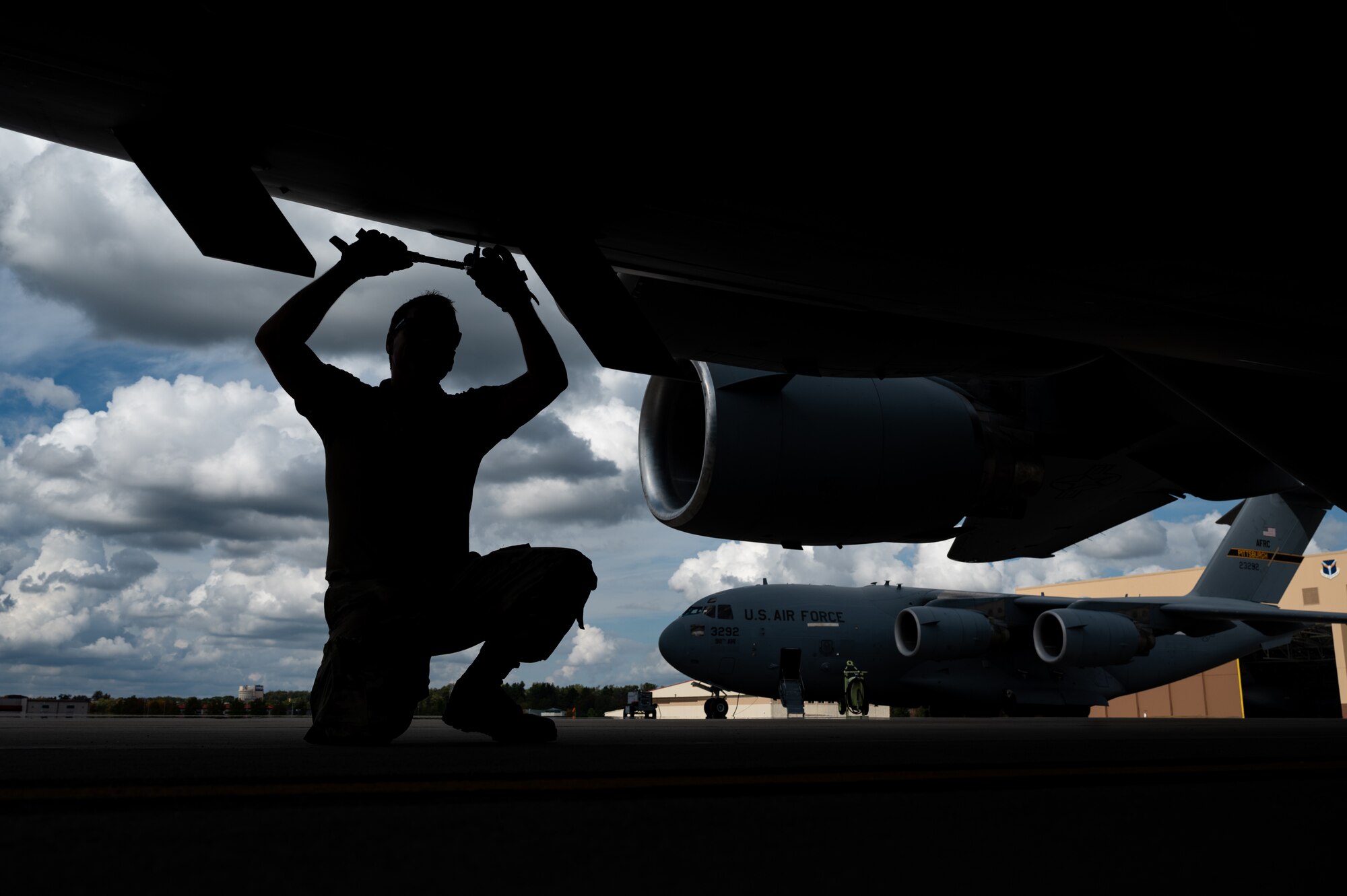 A kneeling man is silhouetted against a cloudy sky while holding a wrench and working on a panel under a large aircraft