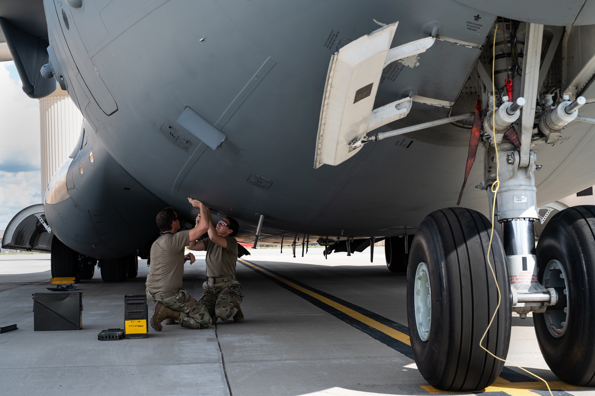 Two Airmen kneel below the underbelly of a large aircraft to work above their heads on a panel