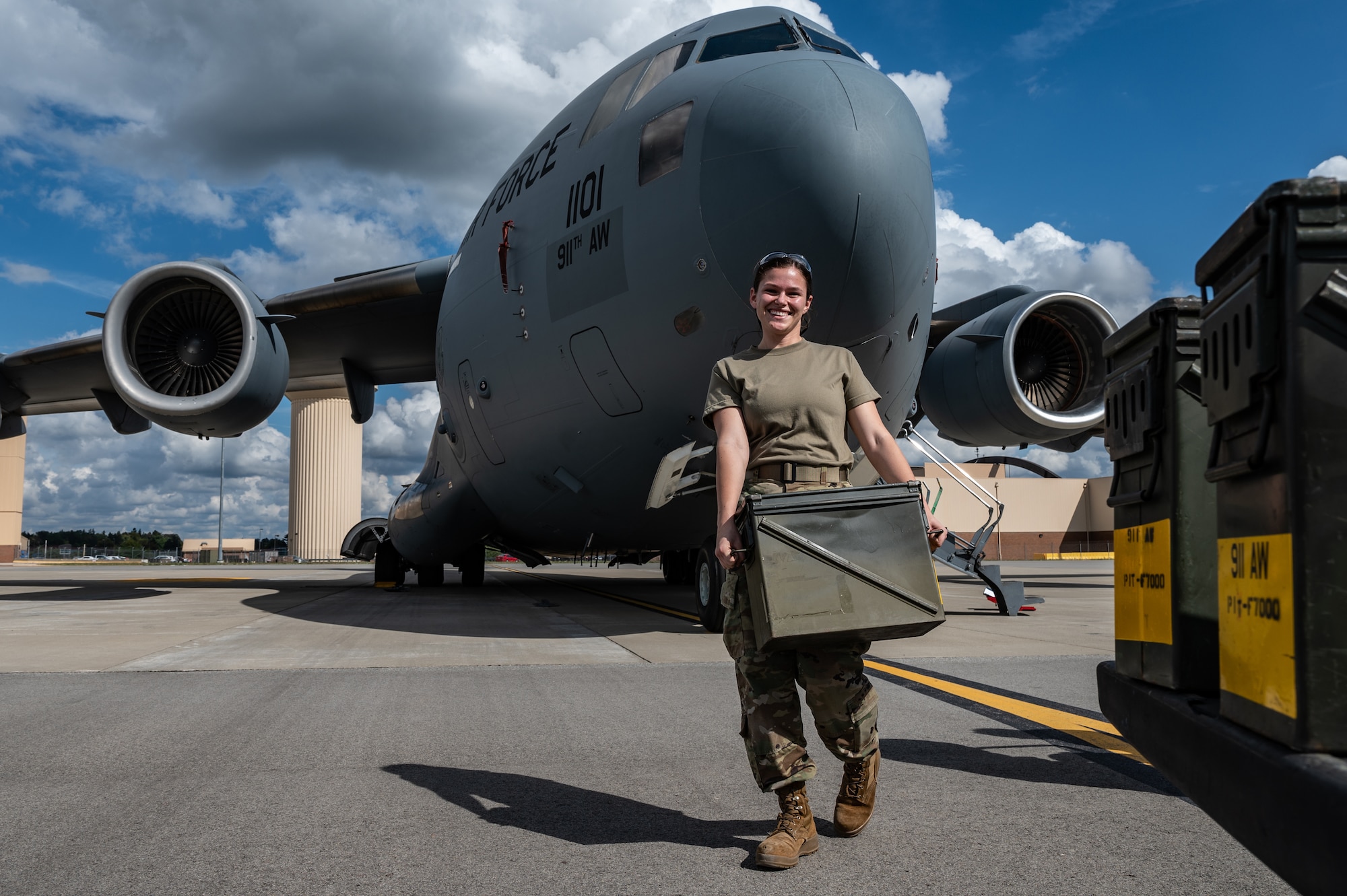 Airman holding a green box smiles while walking in front of a C-17