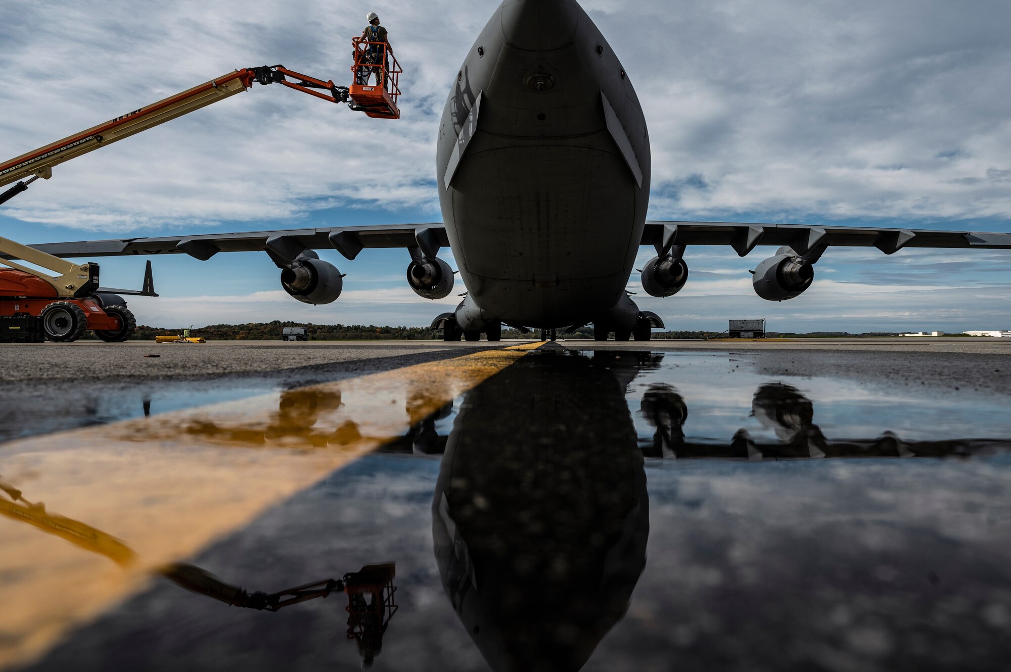 On the left, an orange cherry picker lifts next to a large aircraft, which is reflected in a pool of water