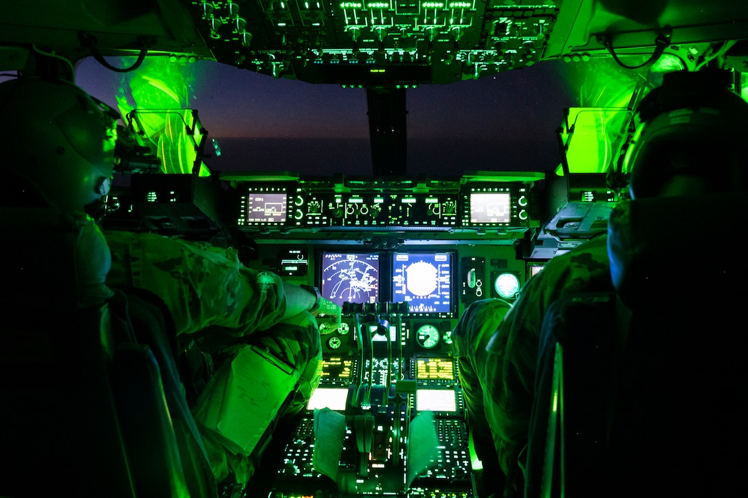 Two pilots sit in a cockpit lit up by green lights.