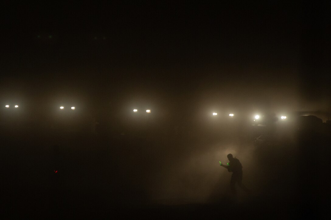 A Marine guides a vehicle through fog at night with headlights of other vehicles in the background.