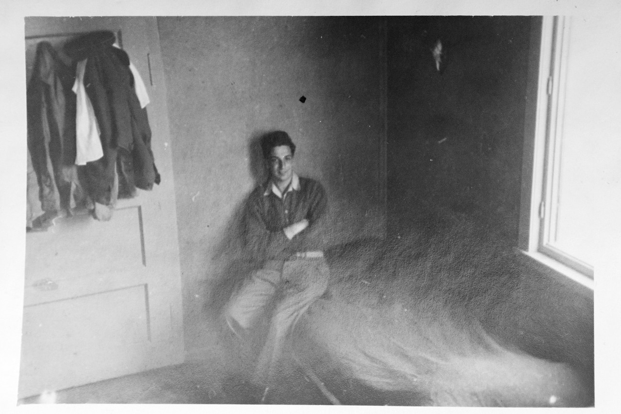 A soldier sits on the edge of his bed with his arms folded as he poses for a photo.