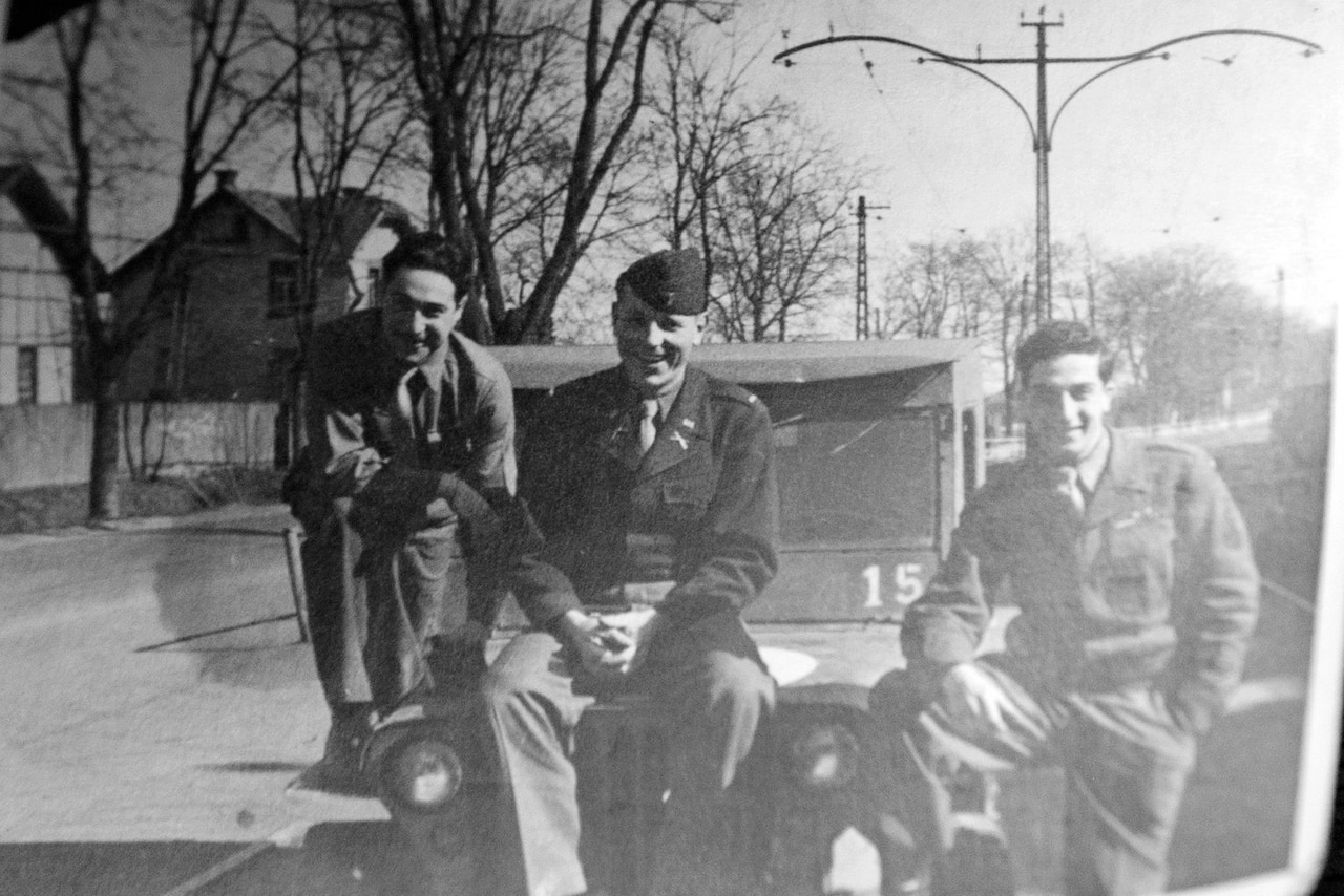 Three soldiers pose for a photo; one is sitting on the hood of a vehicle, another has his foot on the bumper and the third is stooped down beside the vehicle.