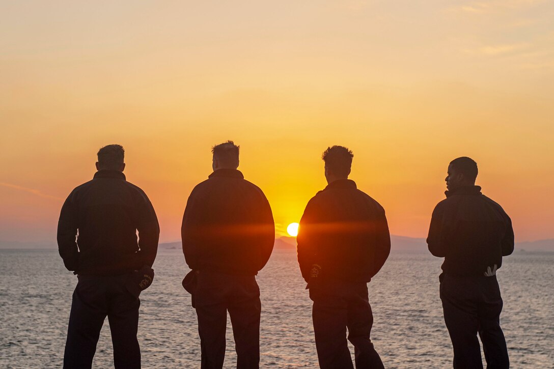 Four sailors stand on the deck of a ship while sun sets on the horizon.