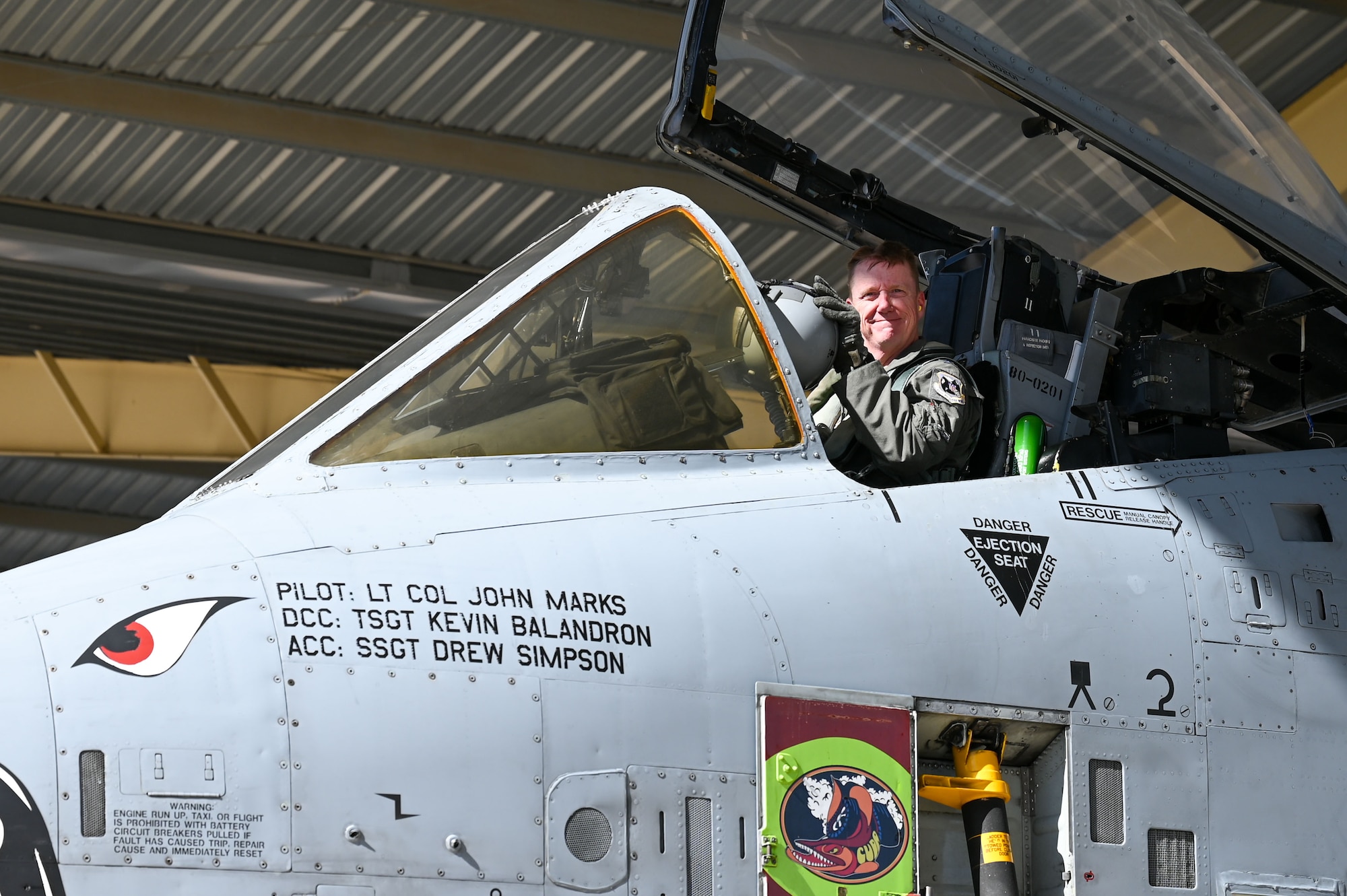 A man smiles from the cockpit of an A-10 Thunderbolt II attack aircraft while holding a helmet.
