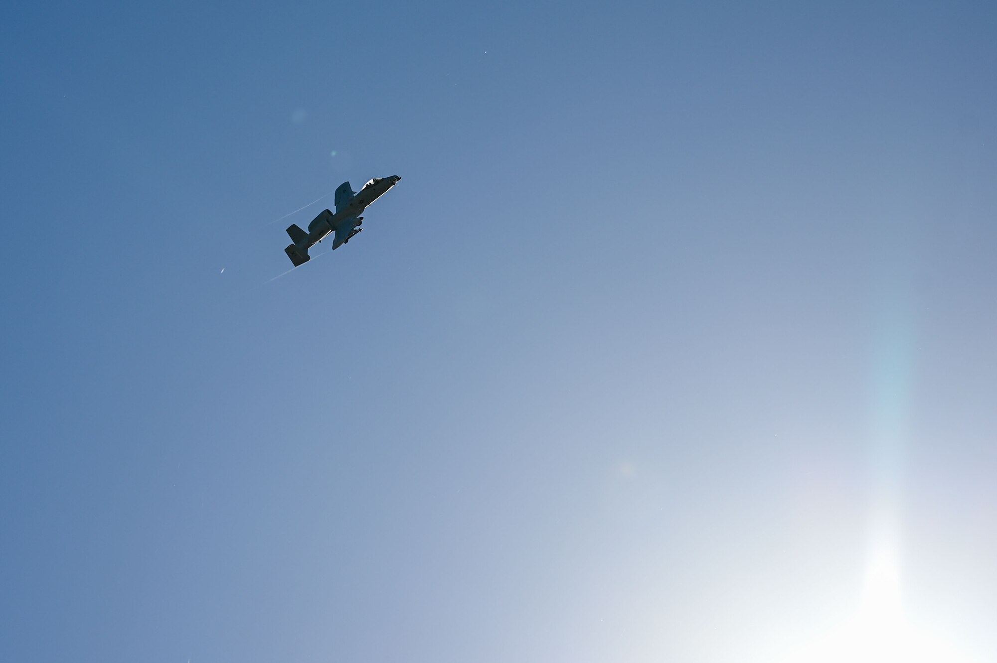 An A-10 Thunderbolt II attack aircraft flies over the sun in a blue sky. Faint vapor trails can be seen coming from its wingtips.