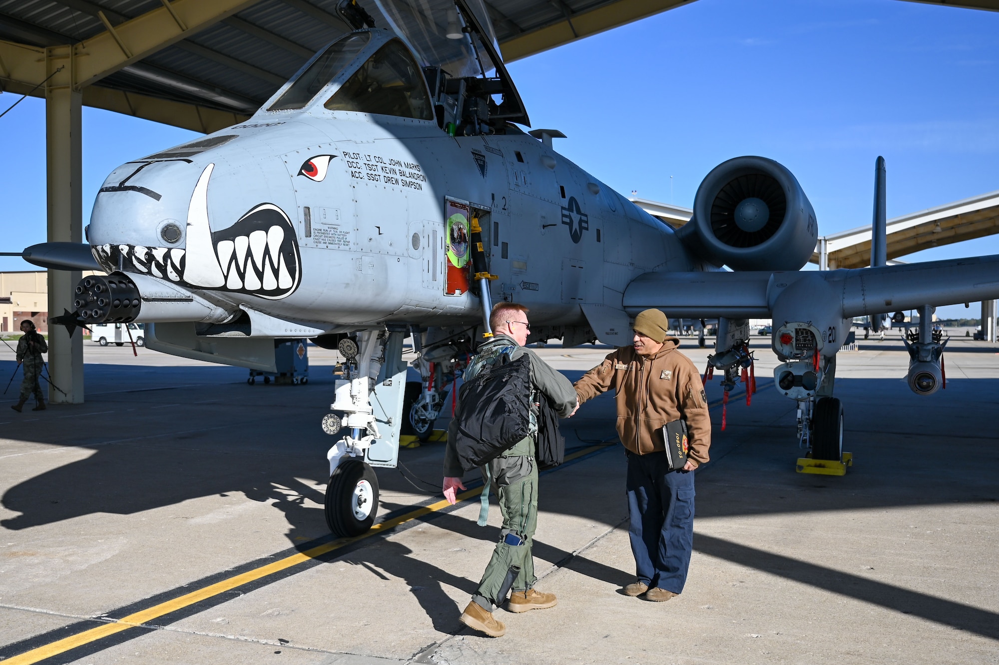A man in a green flight suit carrying a black backpack shakes the hand of a man in a brown coat over blue overalls next to an A-10 Thunderbolt II attack aircraft.
