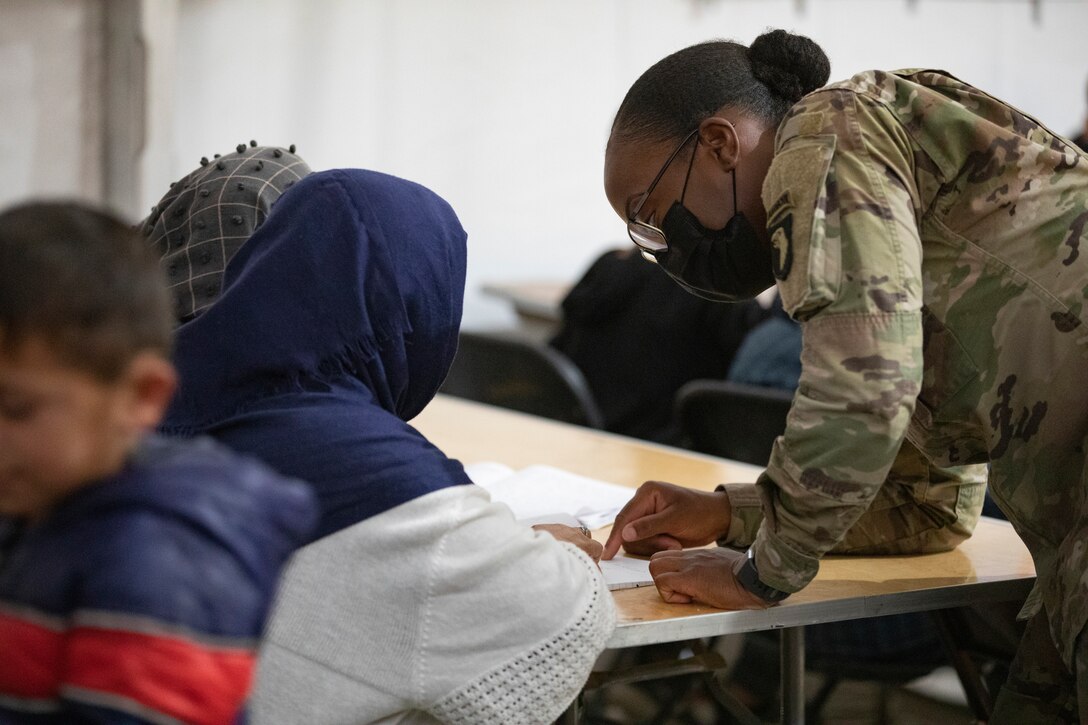 A soldier leans on a table and helps a girl with her school work.