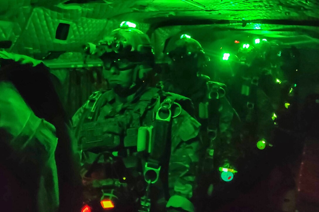 Soldiers wearing protective gear stand in a line on an aircraft illuminated by green light.