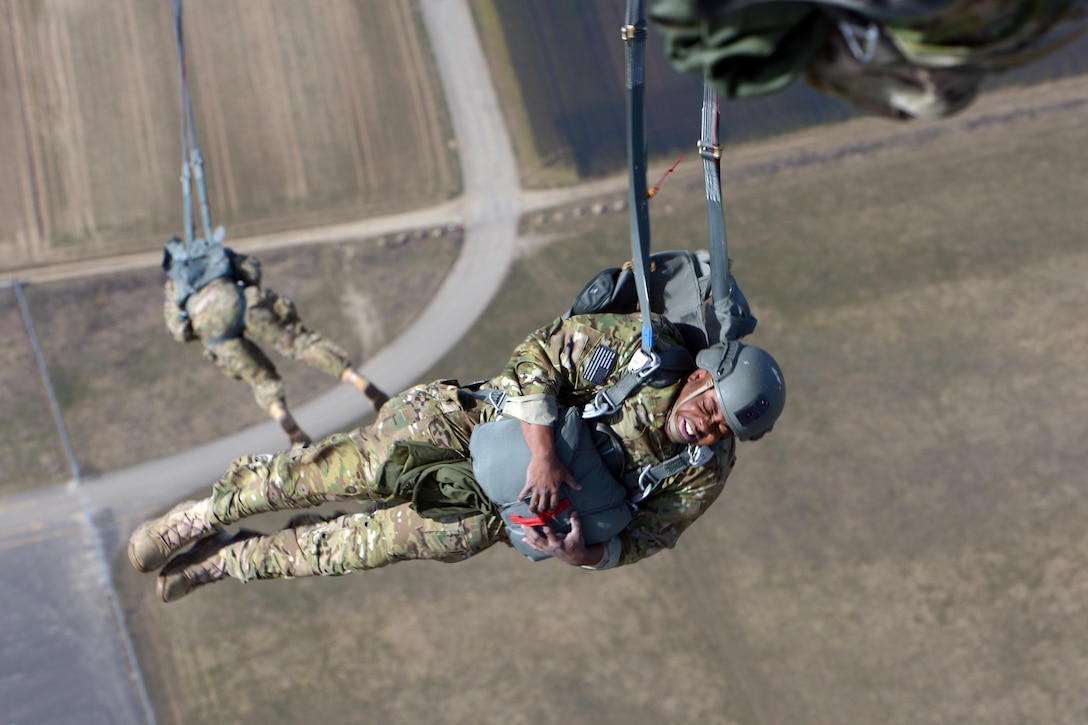 A male soldier grimaces and holds something close to his chest after parachuting from a plane; another soldier floats beneath him.