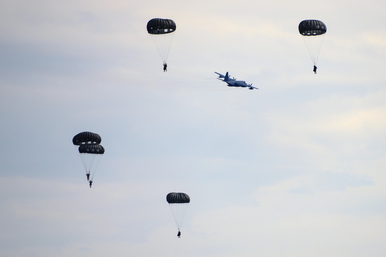 Five soldiers float in the air on parachutes; a plane flies away from them.