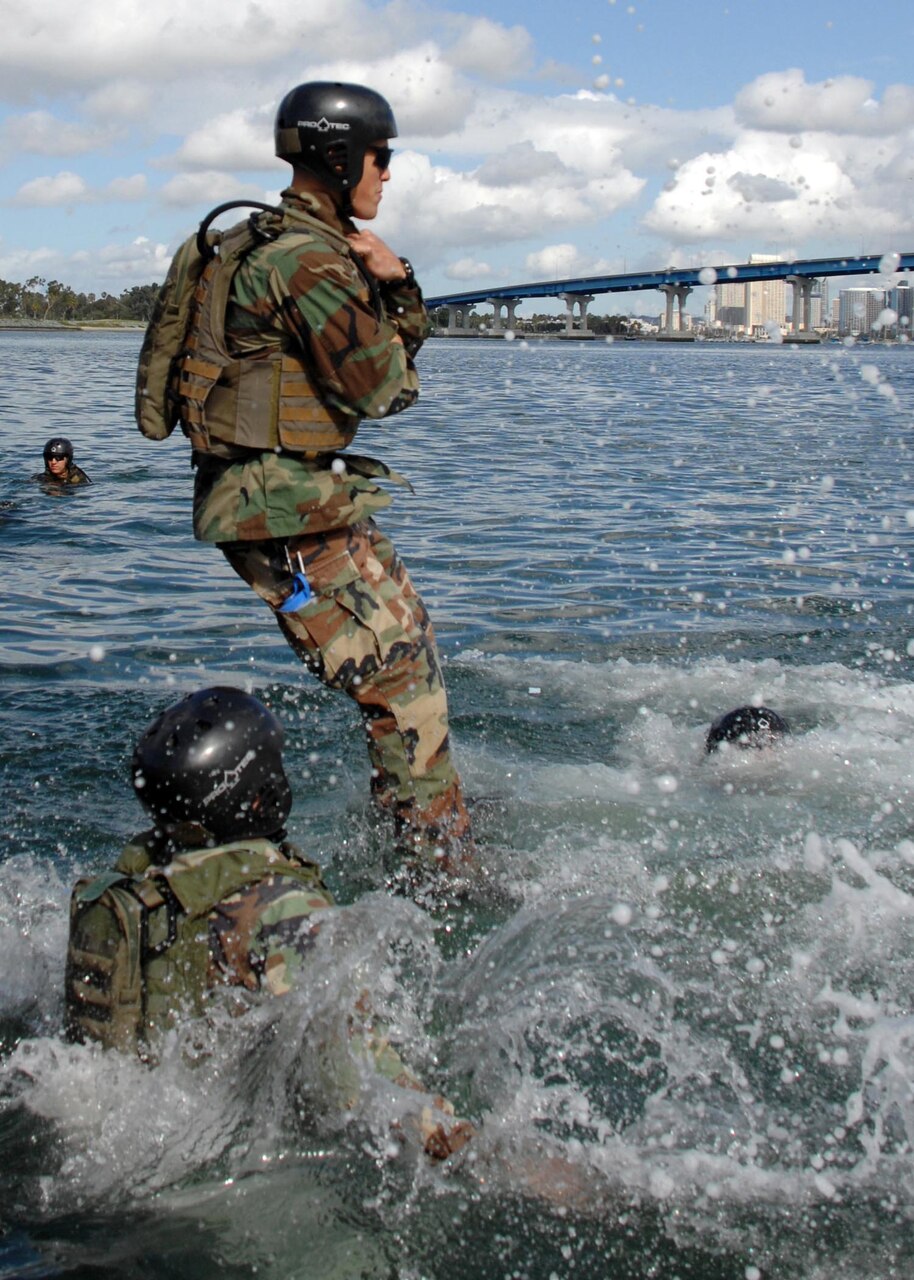 A uniformed sailor jumps into water churning beneath him; the heads of three other service members bob in the water. A bridge is visible in the background.