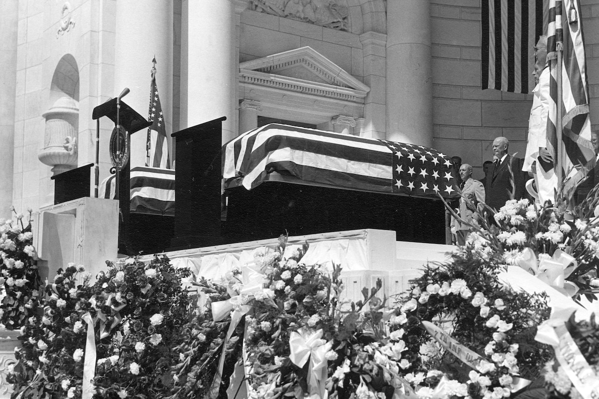 President Eisenhower stands behind two flag-draped coffins on a raised platform with flowered wreaths below.