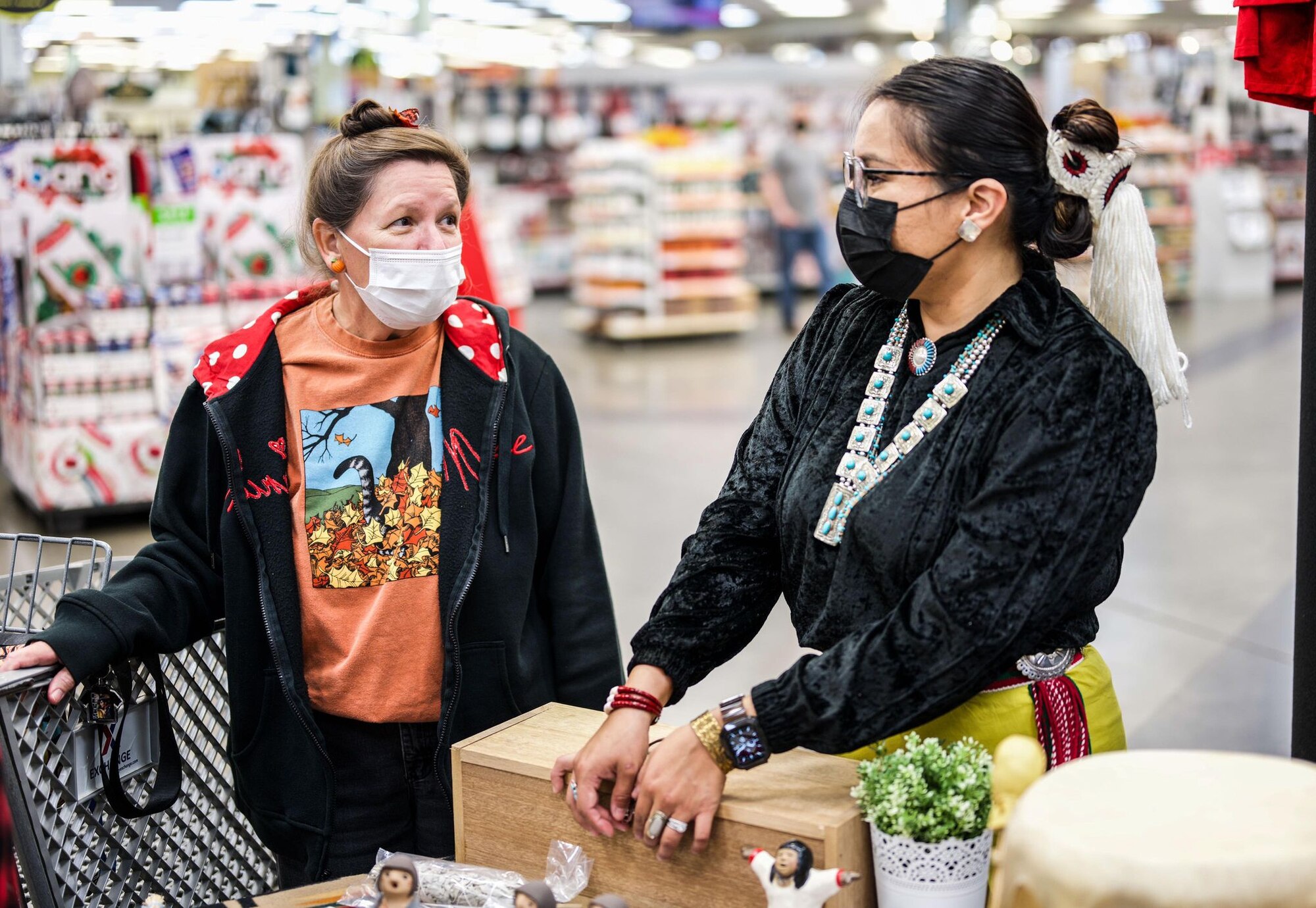 A woman in Native American regalia speaks with a woman in a black hoodie inside a department store setting