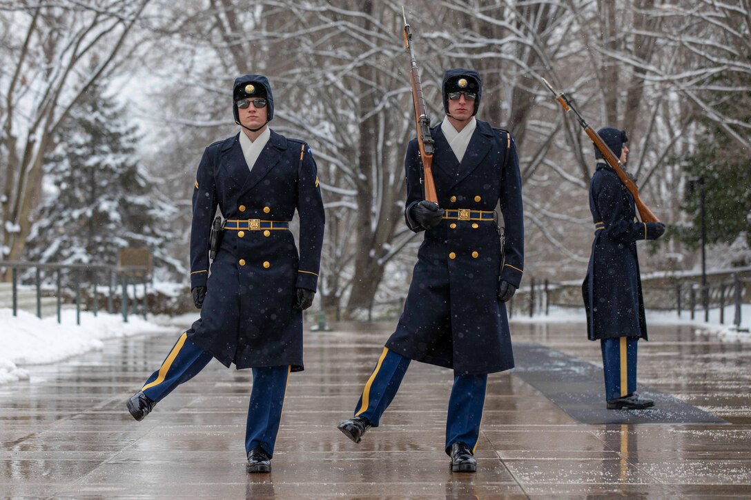 Two soldiers walk move with rifles while another stands guard in winter weather.
