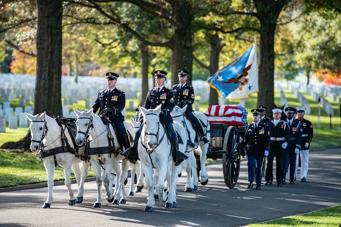 Service members on white horses lead a wagon with a casket on it. Other service members walk beside it.
