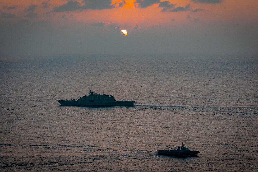 A large and small ship sail in the ocean while the sun sites in the sky overhead.