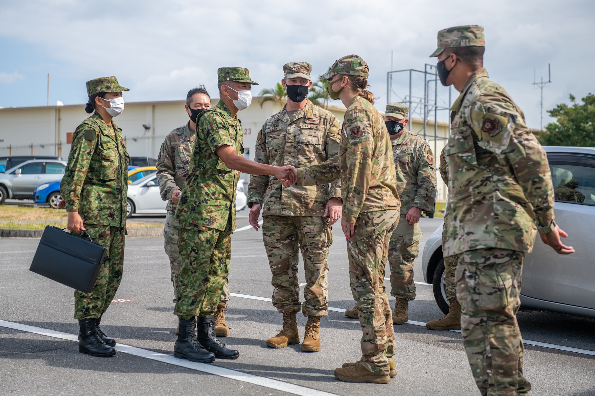 A group of military members stand around a Japanese military member shaking hands with an American military member