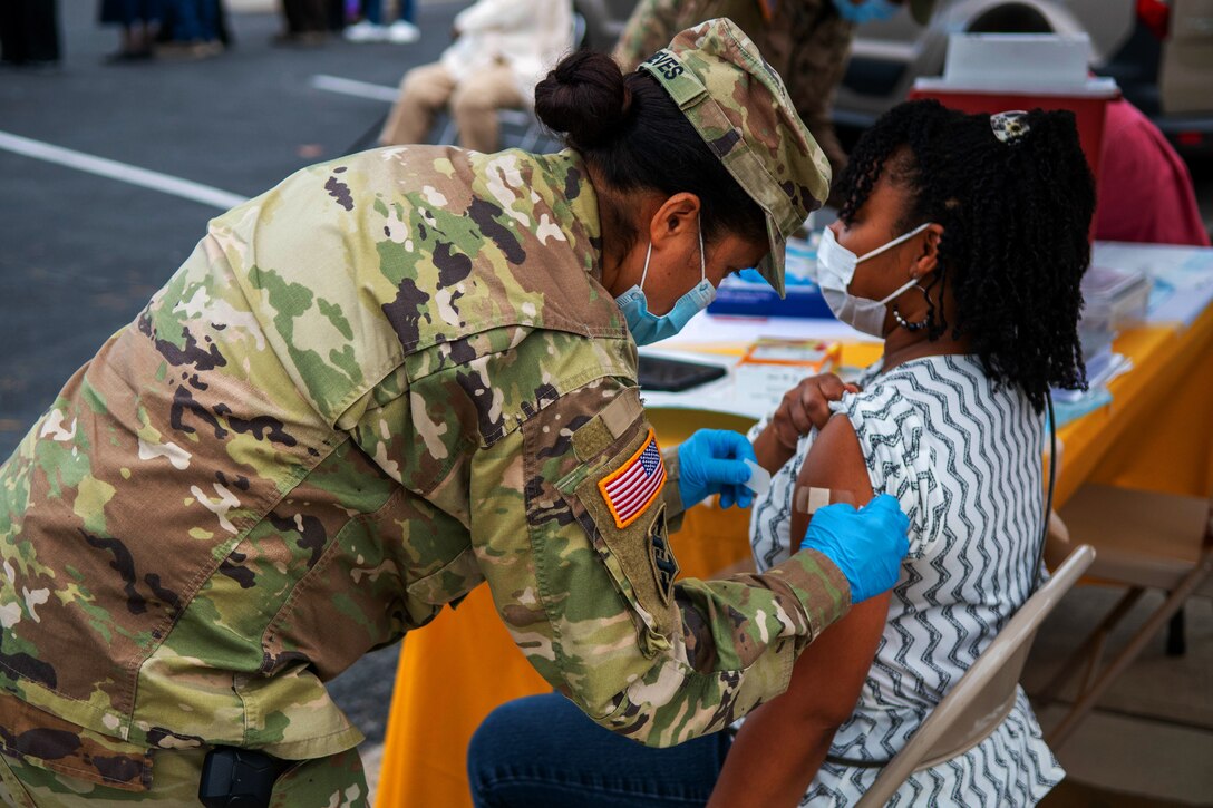A service member wearing a face mask, gloves and a cover leans over to put a bandage on the arm of a Maryland resident.