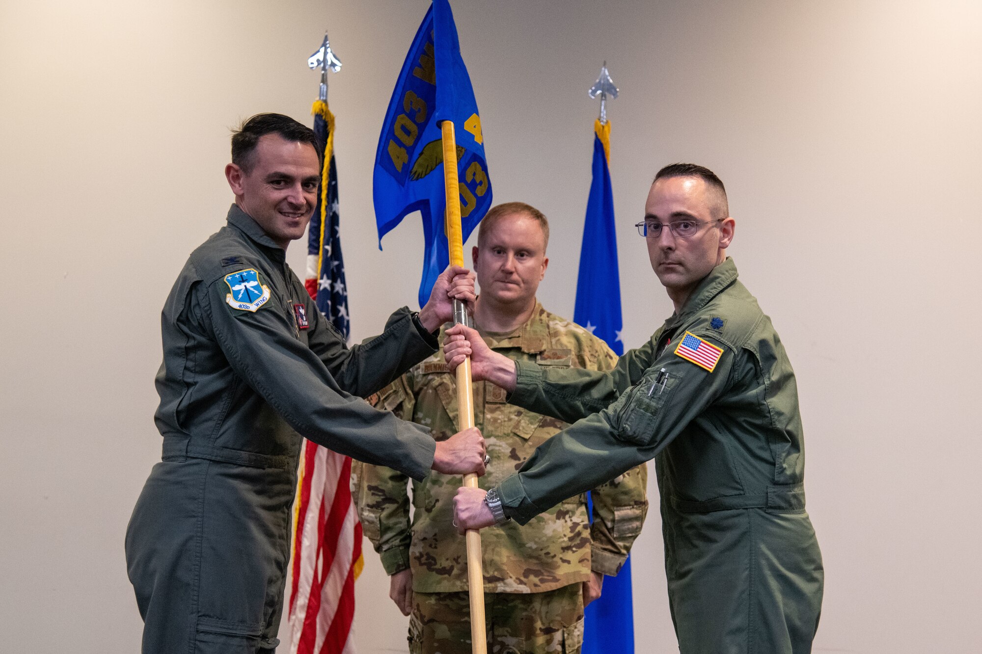 Two officers in flight suits hold a guidon staff. Guidon bearer stands at attention in the center behind them flanked by two flags.