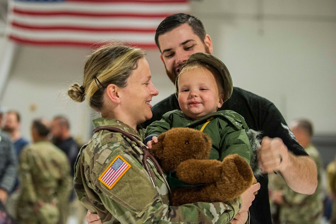 A soldier holds a toy bear while a man and baby smile.