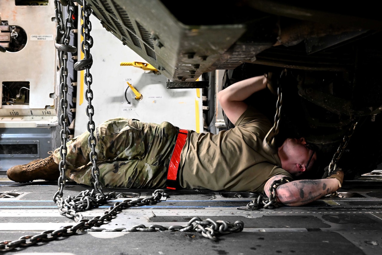 An airman lies on underneath a military vehicle.