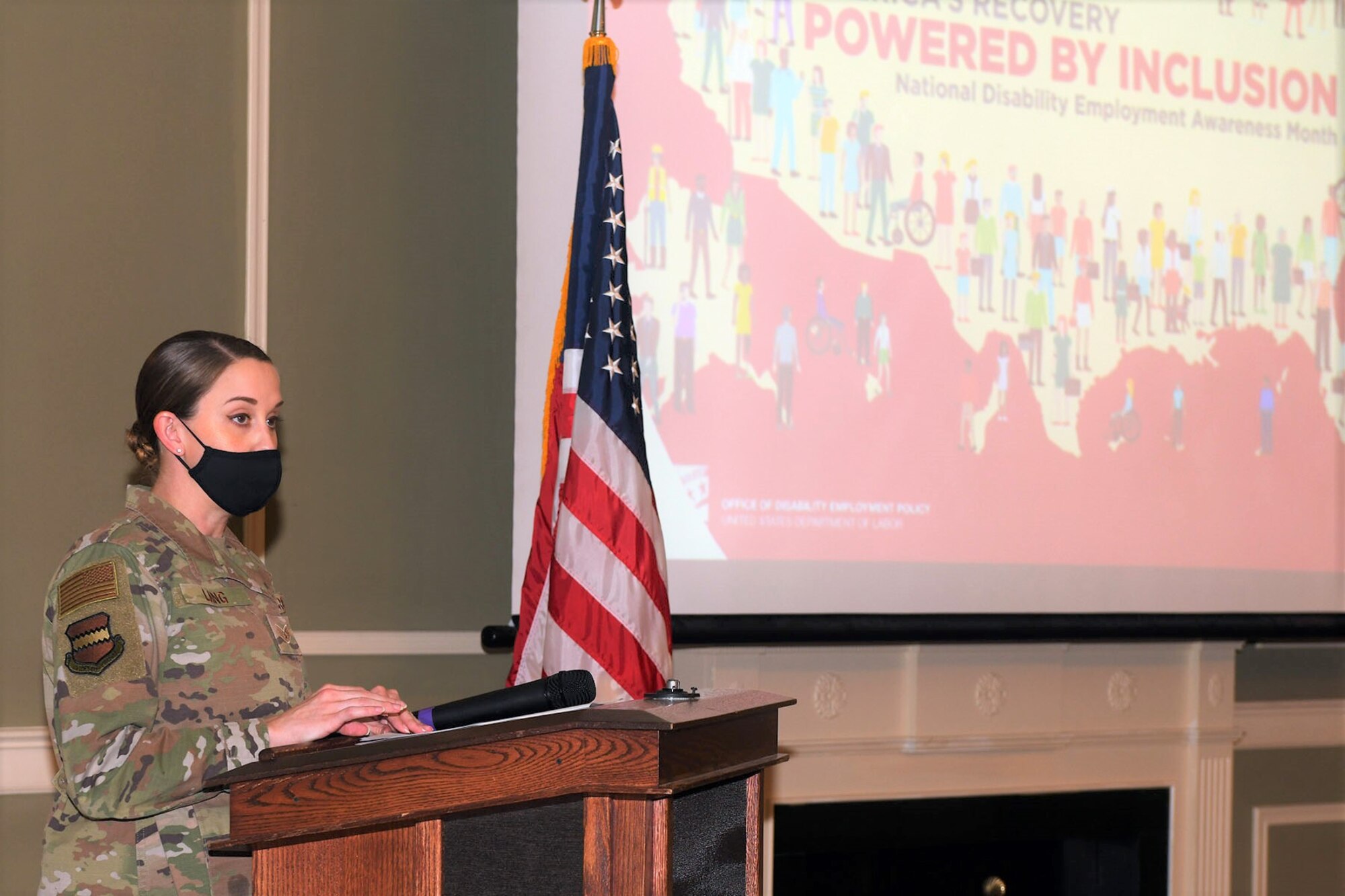 Female Air Force member in camouflauge uniform speaks at a podium.