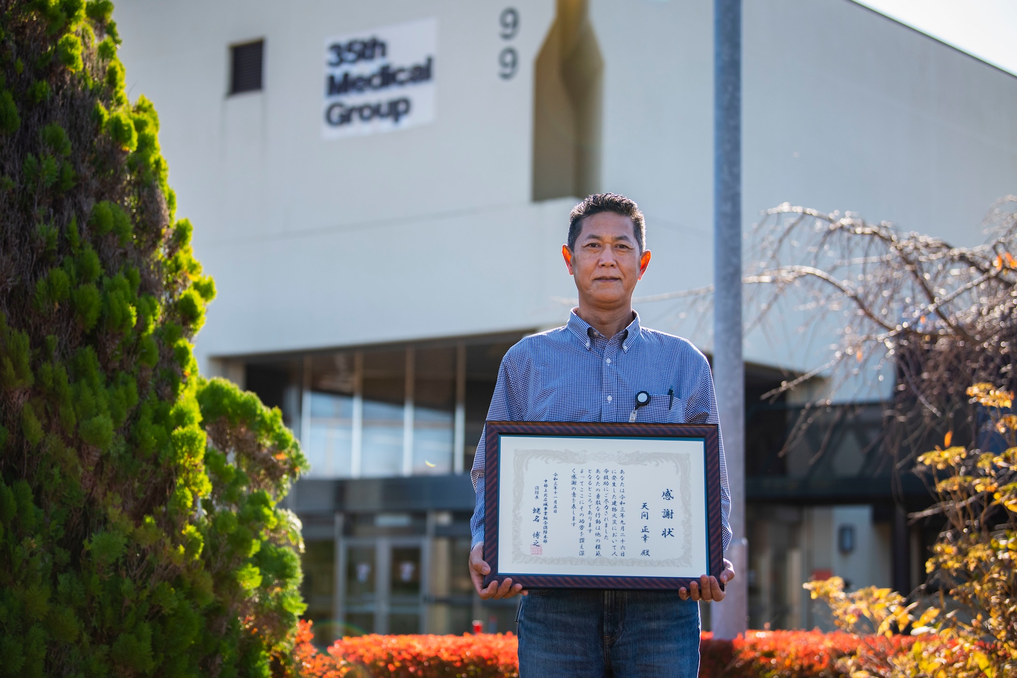 Man stands in front of medical building holding up a framed document.