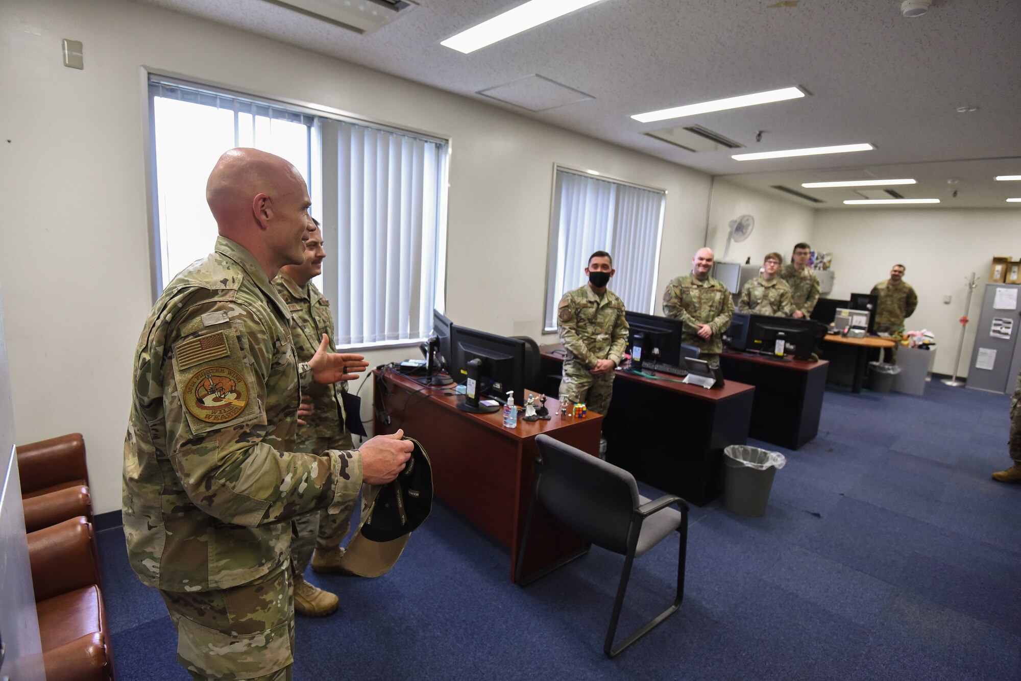 Military members stand in front of their desks while another military member speaks to them.