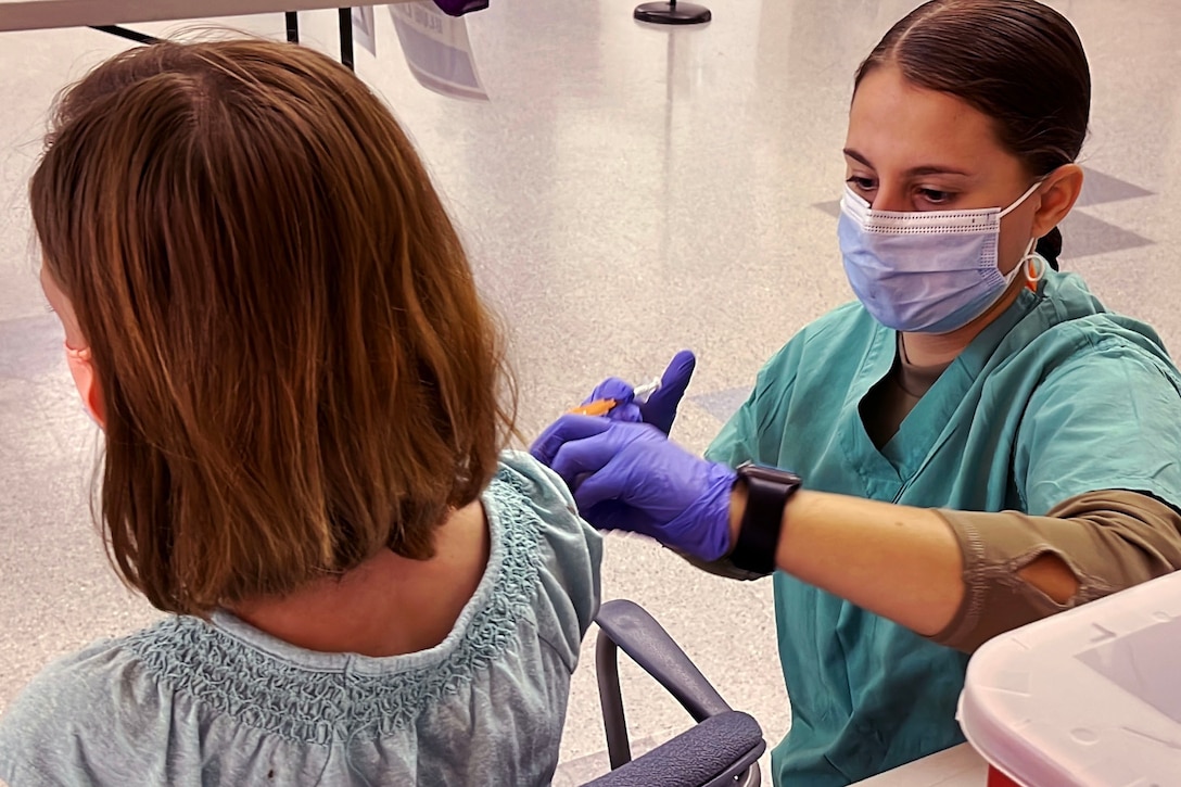 A female medical tech wearing a face mask and gloves kneels down while holding a syringe to a child who is seated.