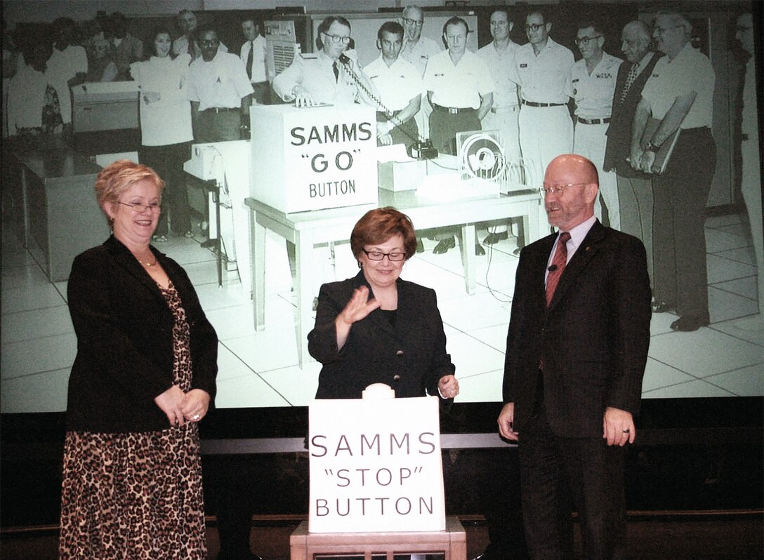 Two women and one man stand in front of a slide show presentation and behind a podium that reads, "SAMMS 'stop' button"