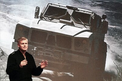 Navy Vice Admiral (male) poses in front of a photo of an MRAP.