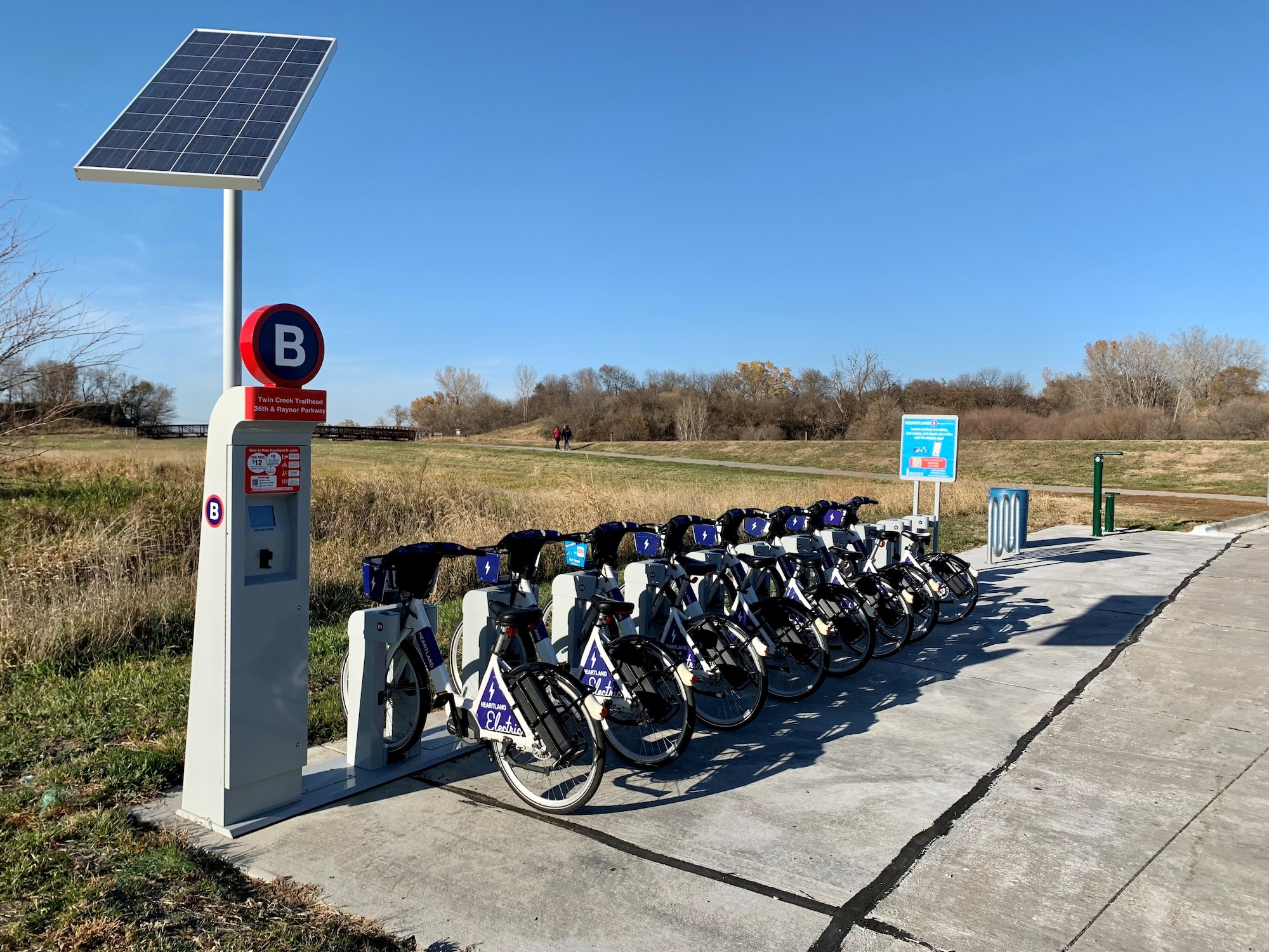 E-bike station with e-bikes in rack and kiosk for rental in front of a trail area