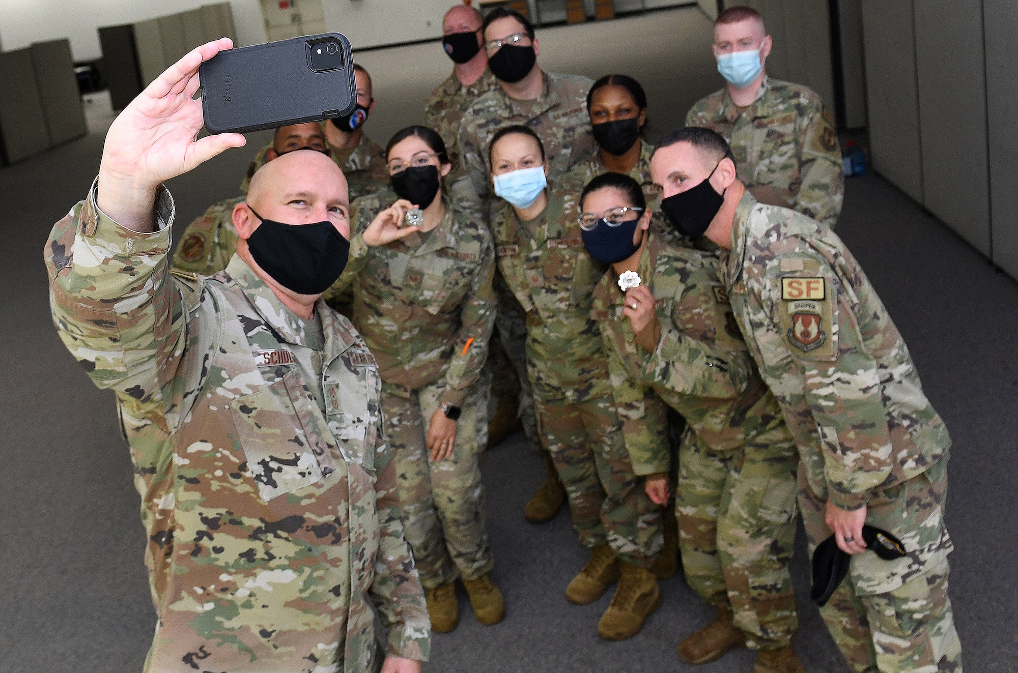 Photo shows a man holding a cell phone up with a group of Airmen behind him all posing for the camera.