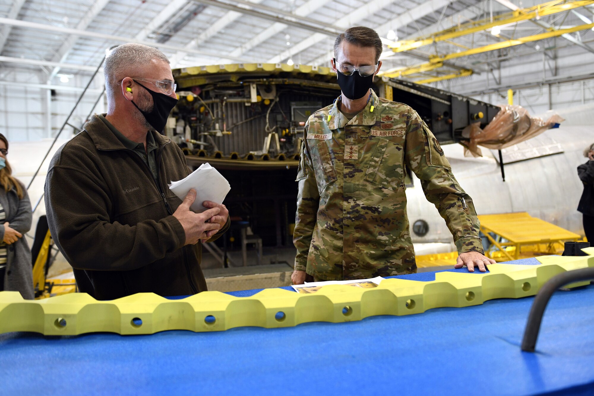 Photo shows two individuals looking at yellow aircraft part on table.