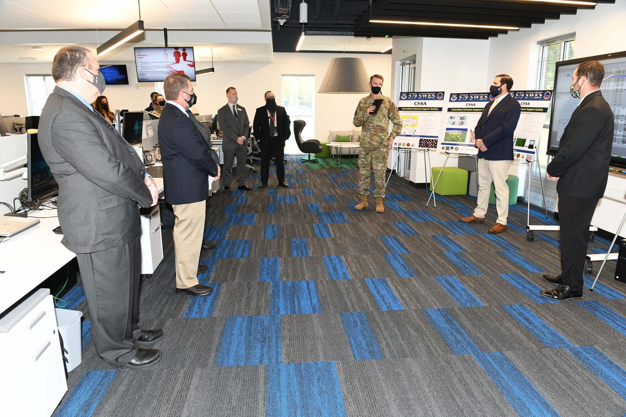 Photo shows people standing in large room with desks talking.