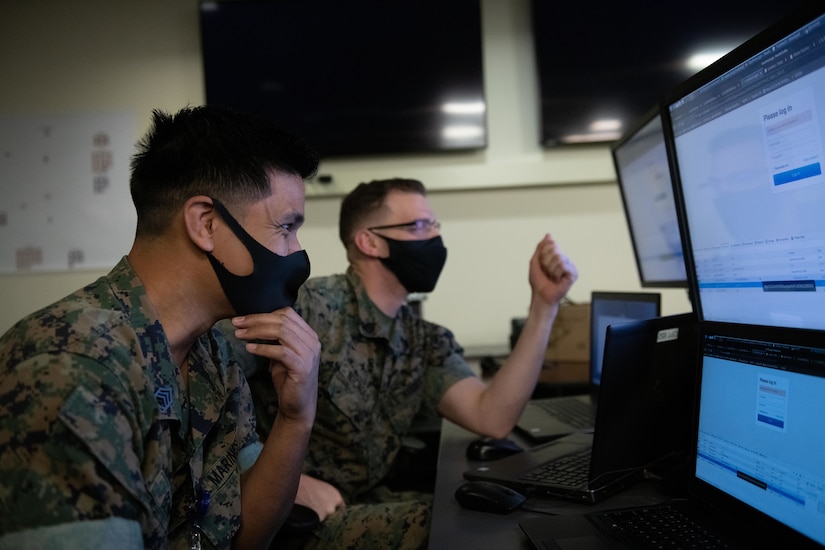 Two Marines wearing masks look at computer screens.