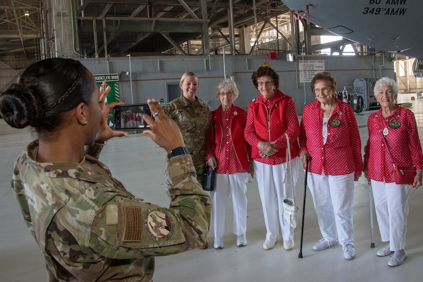 An airman poses for a photo with four Rosie the Riveters in an airplane hangar.