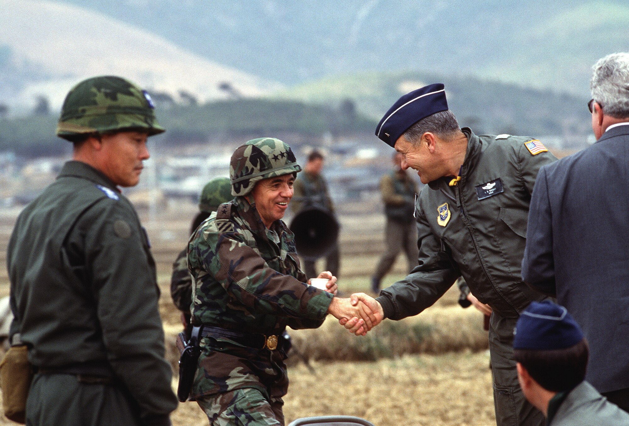 High ranking military men shake hands in a field surrounded by other military members.