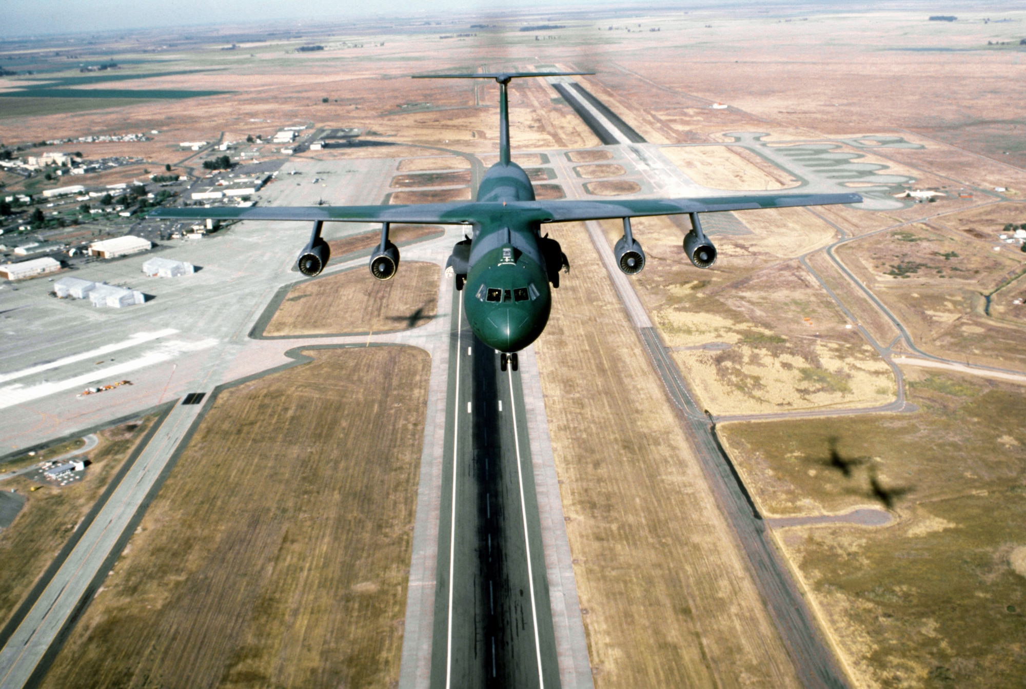 A large camouflaged military aircraft in the sky over a runway.