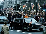 Two people are outside the top window in a car waving to people as it drives through a parade with the streets lined with a lot of people.