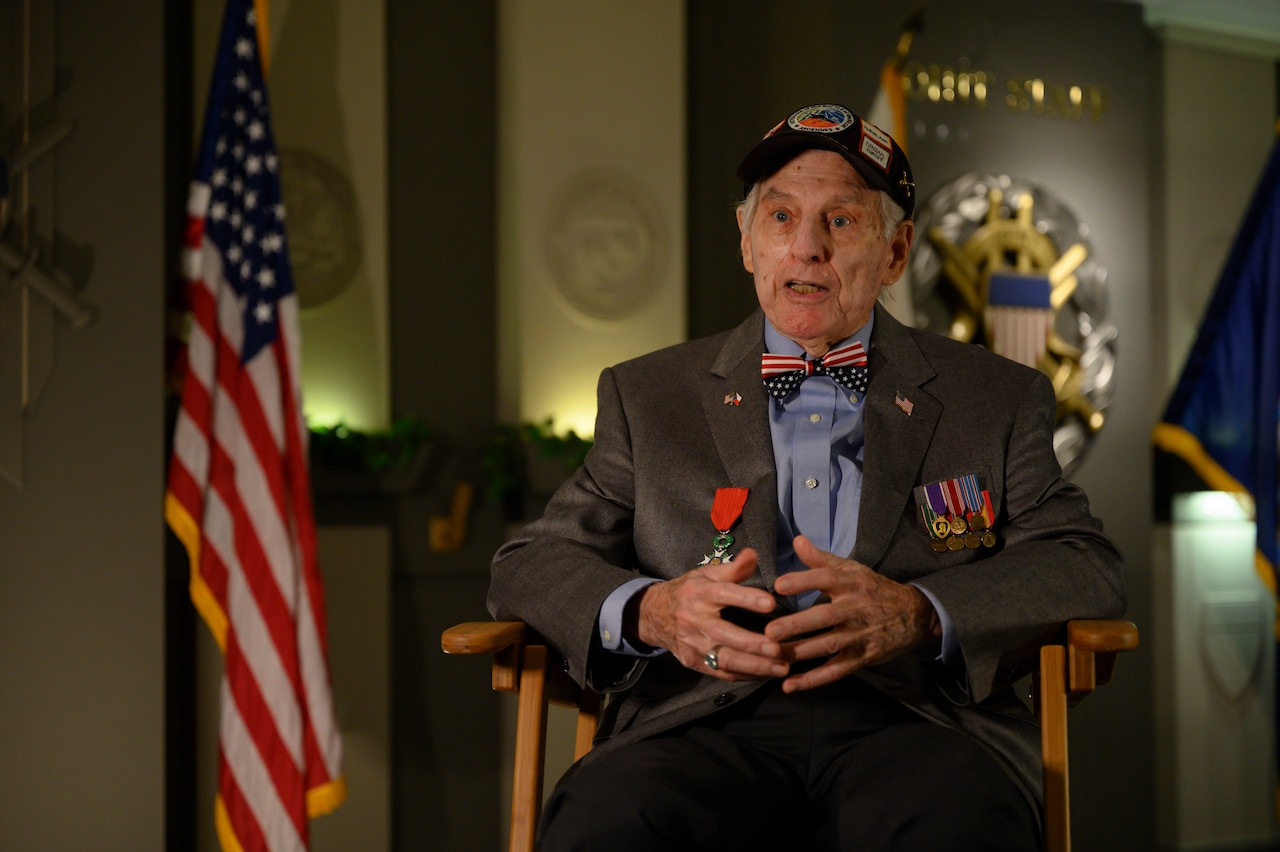 An elderly man appears to be speaking to someone; a U.S. flag is in the foreground.