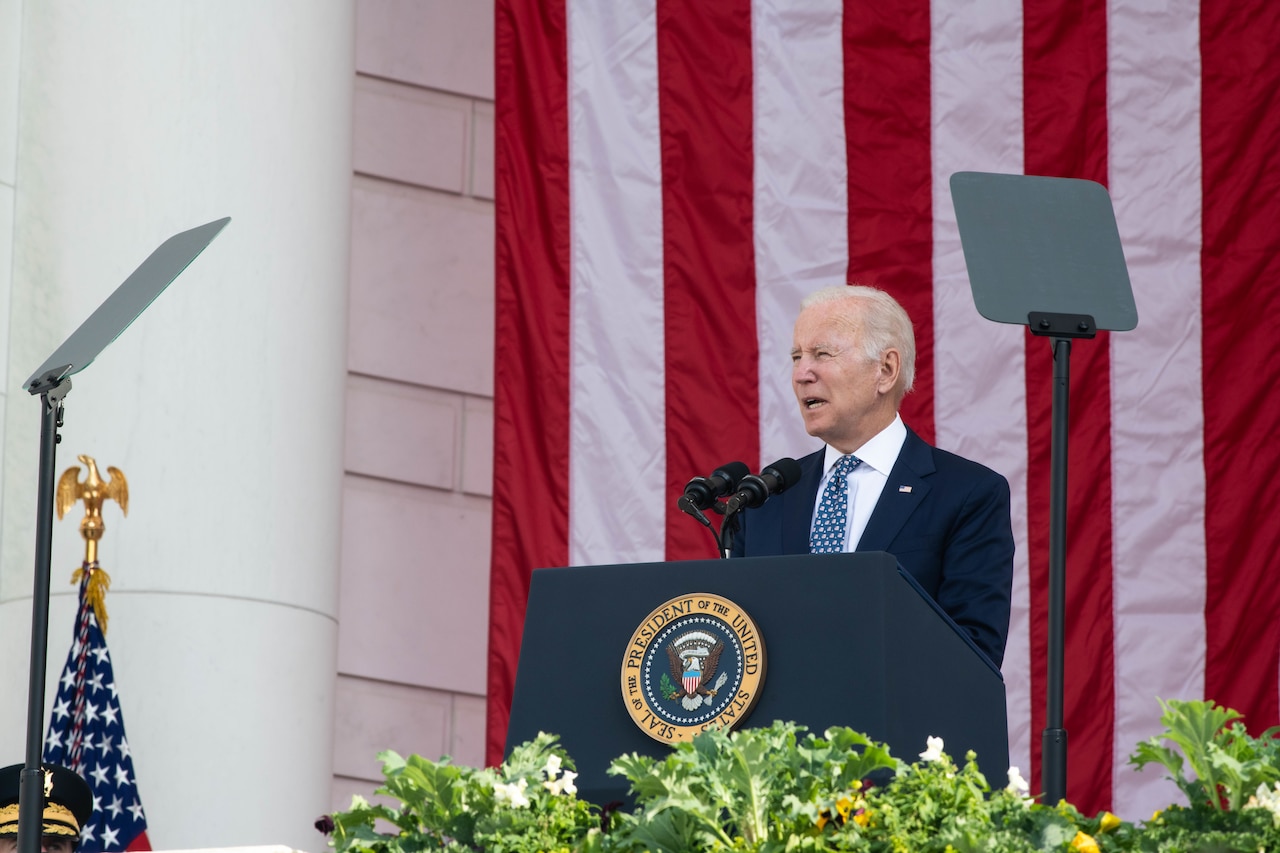 A man stands at a lectern and speaks into a microphone; behind him is a large flag.