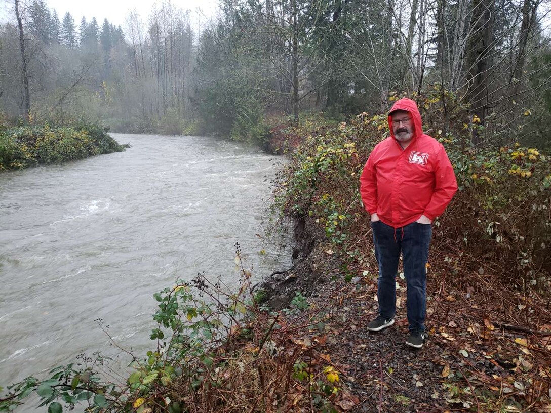 Photo taken of John McAvoy, mechanical engineer with the Seattle District, standing at the bank of the Wallace River in the Snohomish River Basin, to show the rise of the river bank and the degree of bank erosion