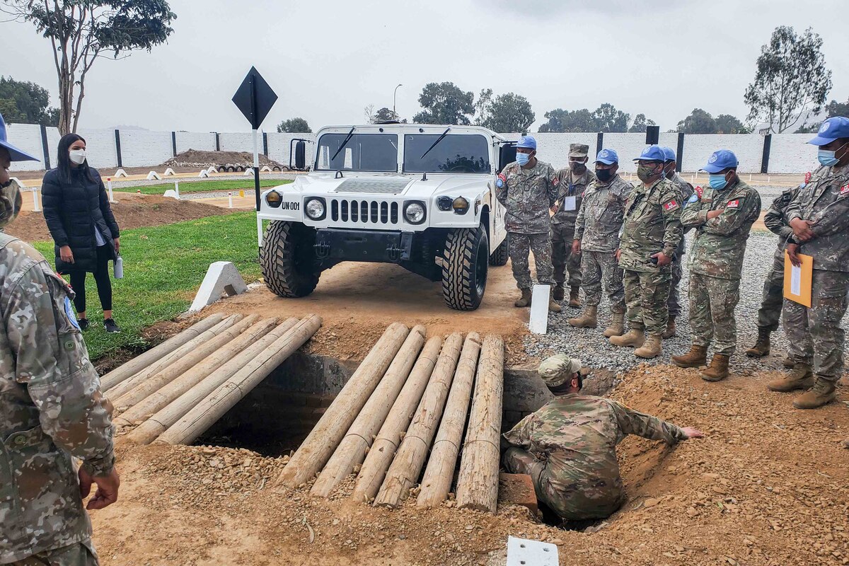 A military vehicle is positioned in front of a large hole; numerous poles are across the top of the hole as service members watch.